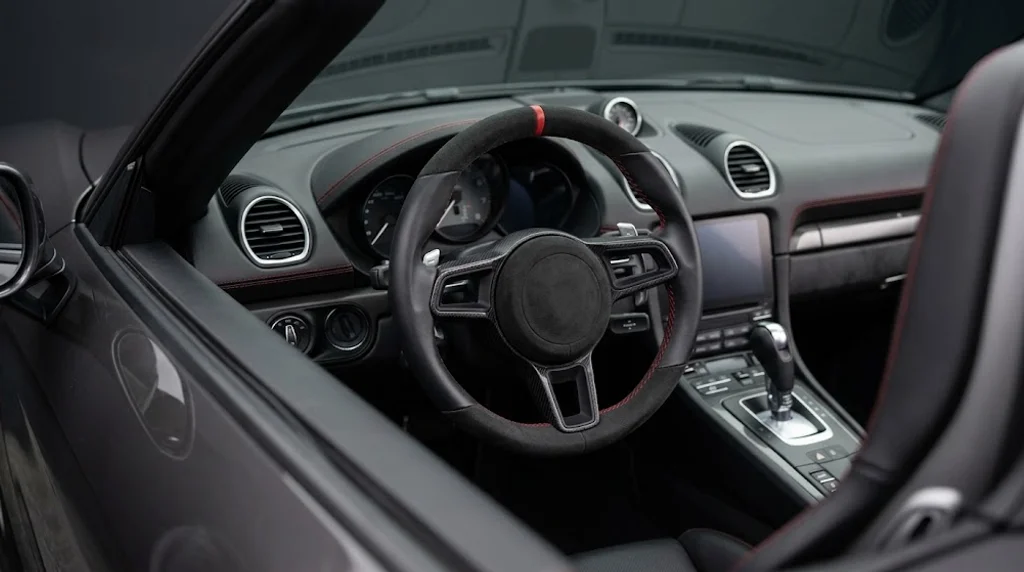 Interior view of a CASSERON custom steering wheel for Porsche 718 Boxster, with black Alcantara rim, matte carbon fiber inserts, guards red stitching and aluminium paddle shifters