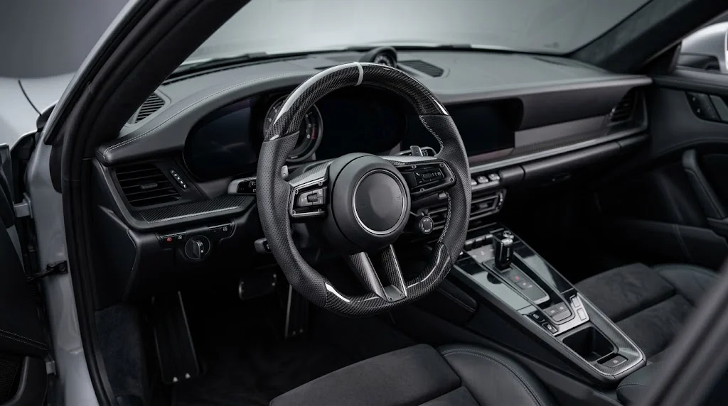 Interior view of a CASSERON custom Porsche steering wheel with gloss carbon fiber on the upper rim and matte carbon fiber on the lower rim, perforated black leather grips and silver contrast stitching